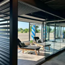 Nowoczesna, przeszklona przestrzeń z leżankami, osłonięta metalową konstrukcją z regulowanymi lamelami, widok na zewnątrz przez duże okna z dekoracją balonową.