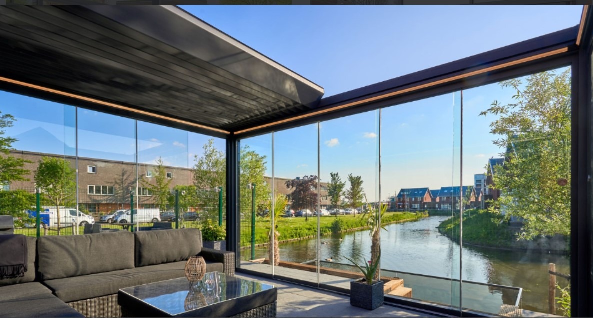 Nowoczesna, przeszklona konstrukcja z metalową ramą, częściowo zadaszona, z widokiem na rzekę i zabudowania mieszkalne w tle. Wewnątrz znajduje się ciemnoszara sofa i stolik kawowy.