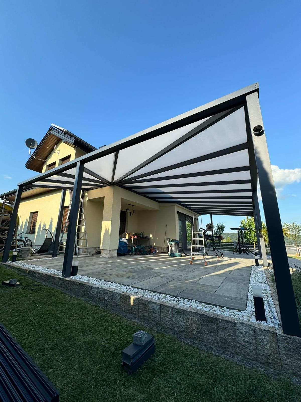 Nowoczesna, czarna konstrukcja z zadaszeniem na tarasie, z widocznymi elementami montażowymi i narzędziami, na tle domu jednorodzinnego i błękitnego nieba.