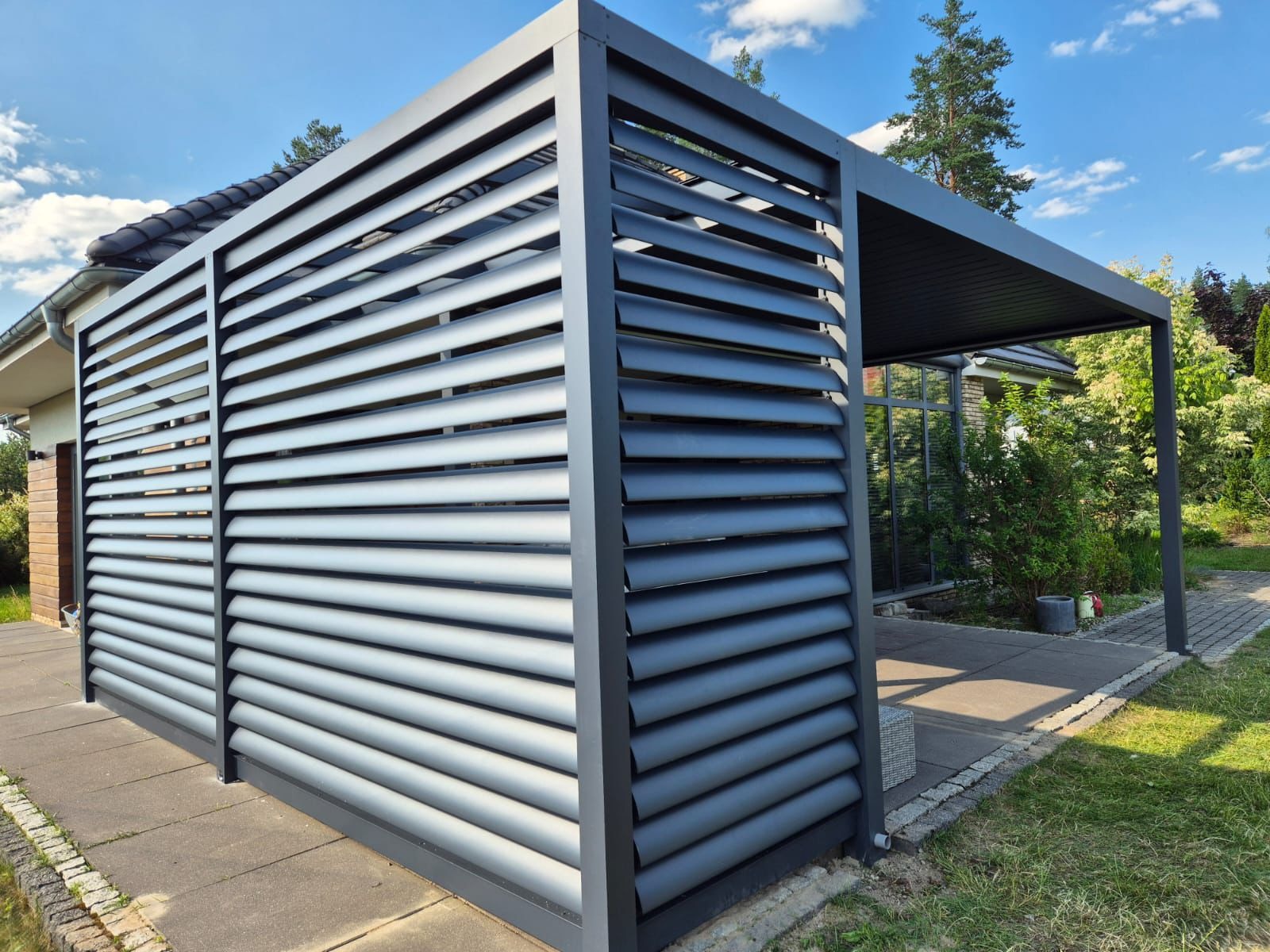 Nowoczesna, szara pergola z metalowymi lamelami, przylegająca do domu z widocznym fragmentem dachu i ogrodu w tle, oświetlona jasnym, popołudniowym słońcem.