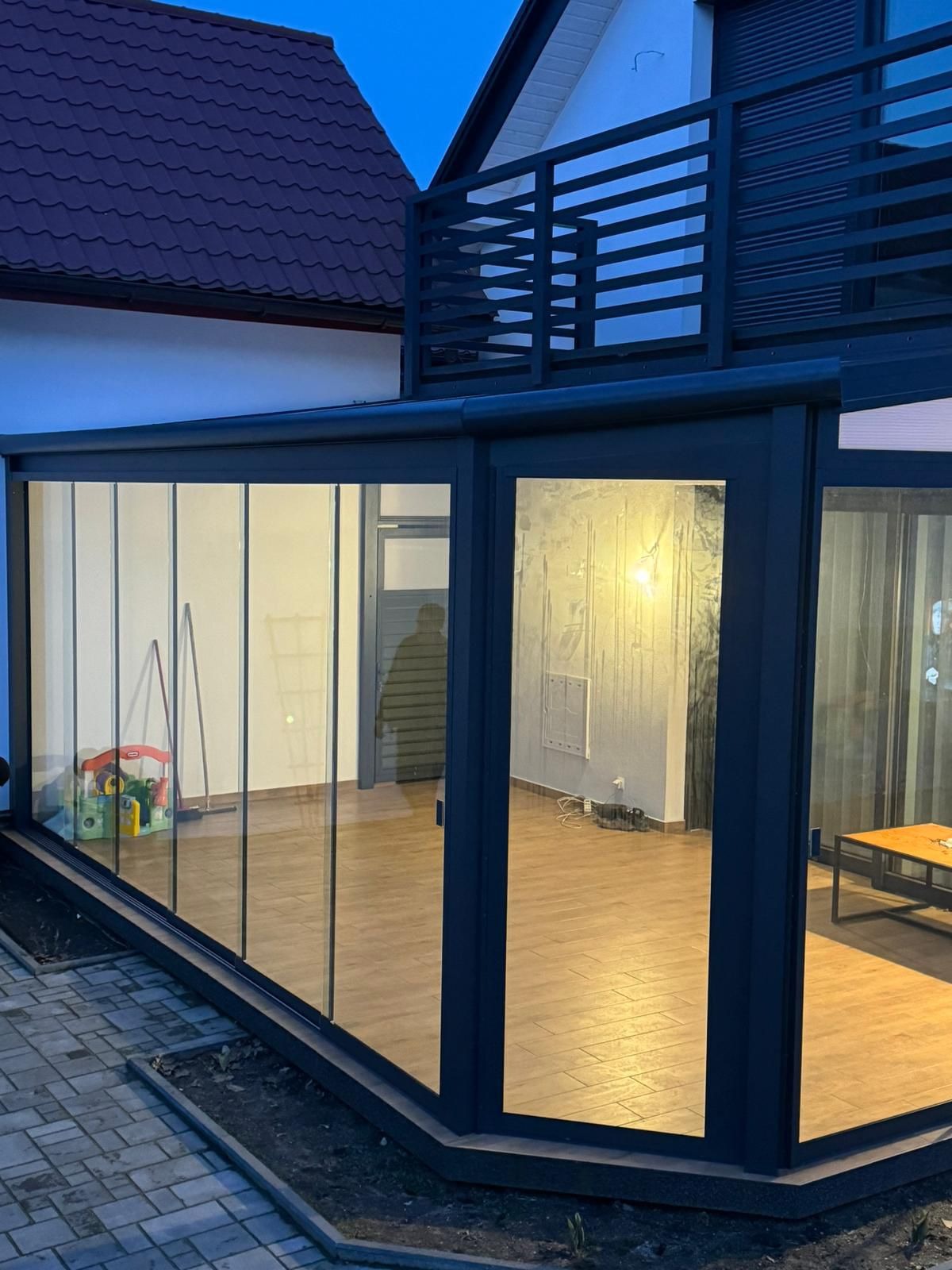 Nowoczesna, przeszklona weranda z ciemną ramą, widok z zewnątrz na drewnianą podłogę, zabawki dziecięce i sylwetkę osoby wewnątrz, z balkonem z metalową balustradą na piętrze domu w tle.