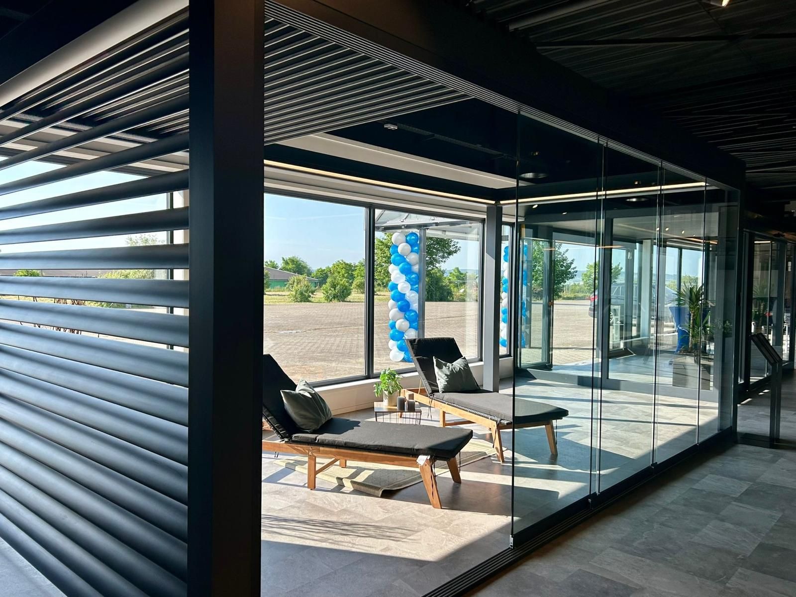 Nowoczesna, przeszklona przestrzeń z leżankami, osłonięta metalową konstrukcją z regulowanymi lamelami, widok na zewnątrz przez duże okna z dekoracją balonową.