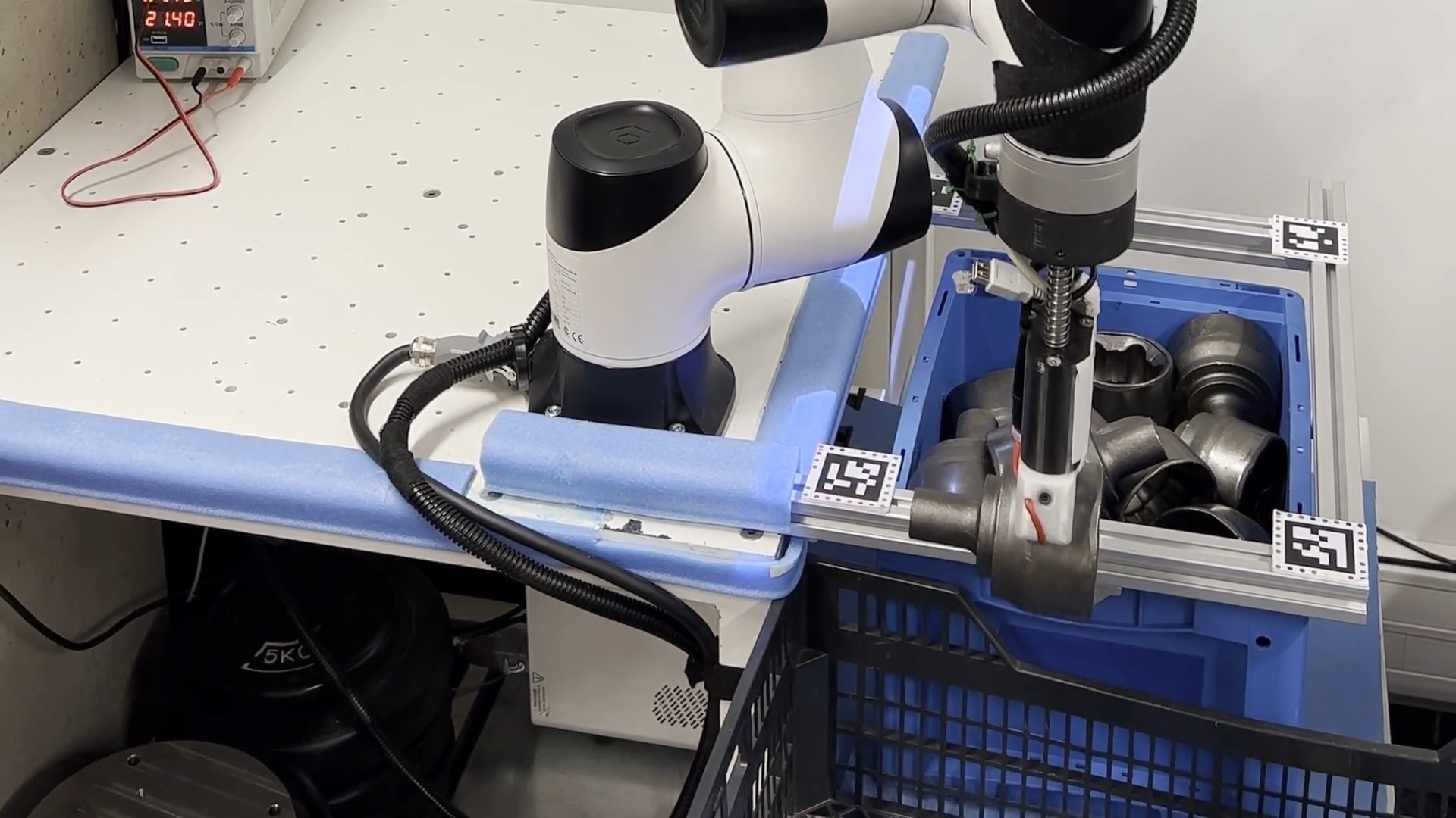 Robot przemysłowy manipuluje metalowymi elementami w niebieskim pojemniku, otoczony aluminiowymi profilami i kodami QR, na tle laboratoryjnego stołu z zasilaczem.