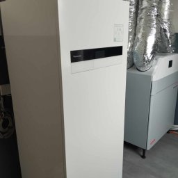 MAR-TECH Zagozda - Biała pompa ciepła Panasonic Aquarea Smart Cloud z widocznymi rurami instalacyjnymi i jednostką wentylacyjną z metalowymi przewodami wentylacyjnymi w tle.