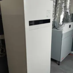 Biała pompa ciepła Panasonic Aquarea Smart Cloud z widocznymi rurami instalacyjnymi i jednostką wentylacyjną z metalowymi przewodami wentylacyjnymi w tle.