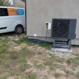 MAR-TECH Zagozda - Czarna pompa ciepła zamontowana na szarym fundamencie obok ściany budynku, z widocznym kablem wchodzącym do skrzynki elektrycznej, na pierwszym planie trawa i częściowo widoczny samochód firmy...