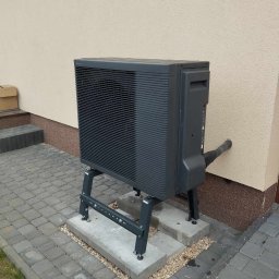 MAR-TECH Zagozda - Szara zewnętrzna jednostka pompy ciepła zamontowana na metalowym stelażu, z rurą przechodzącą przez ścianę budynku, na tle kostki brukowej i elewacji.