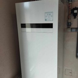 MAR-TECH Zagozda - Biała pompa ciepła Panasonic Aquarea Smart Cloud zainstalowana w pomieszczeniu z widocznymi rurami i zaworami.