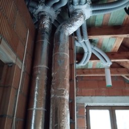 MAR-TECH Zagozda - Dwie pionowe rury owinięte folią aluminiową, połączone z szarymi elastycznymi przewodami wentylacyjnymi i białymi rozdzielaczami w surowym wnętrzu poddasza z ceglanymi ścianami i drewnianą...