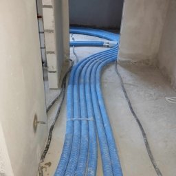 MAR-TECH Zagozda - Niebieskie rury rekuperacji ułożone w rzędach na surowym betonowym podłożu w trakcie montażu w nowym budynku, widoczne przewody elektryczne w peszlach.