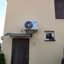 Zewnętrzna jednostka klimatyzacji zamontowana na elewacji domu, widoczna nad wejściem z zadaszeniem i małym oknem z ozdobami.