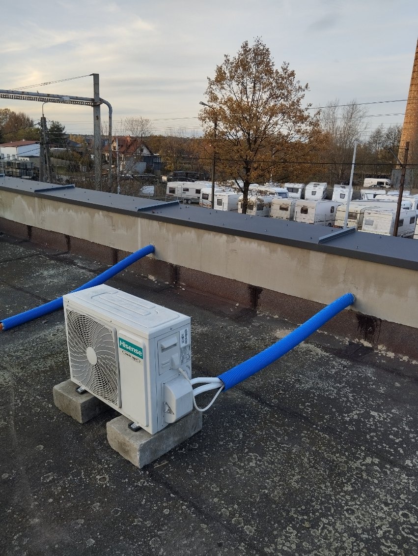 Biała jednostka zewnętrzna klimatyzacji Hisense zamontowana na betonowych bloczkach na płaskim dachu z podłączonymi niebieskimi rurami, w tle widoczne przyczepy kempingowe i komin.