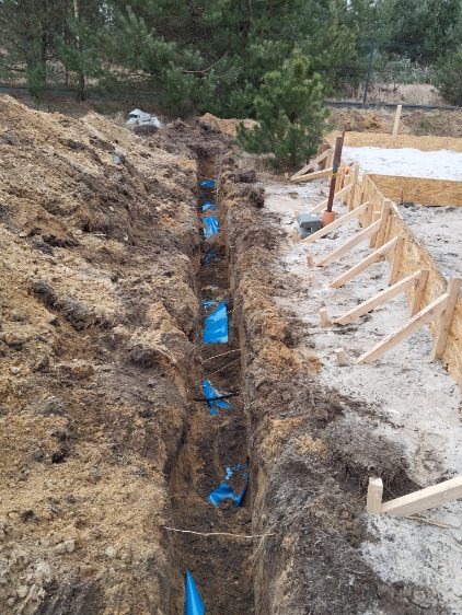 Wykop ziemny z niebieskimi rurami w izolacji, przygotowany pod instalację, obok drewniane szalunki fundamentów.