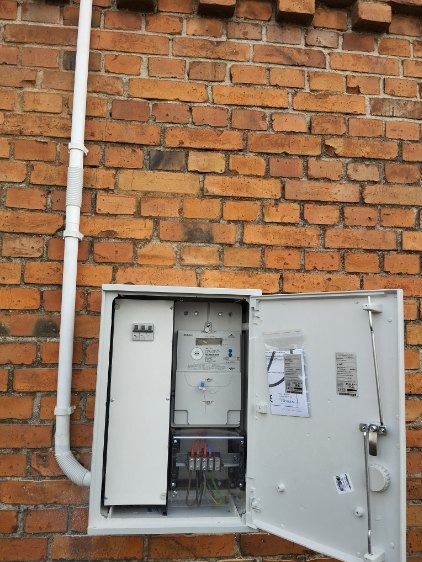 Otwarta skrzynka elektryczna z licznikiem i zabezpieczeniami zamontowana na ceglanej ścianie budynku, widoczna instalacja przewodów wewnątrz skrzynki i rura PCV prowadząca w górę po ścianie.