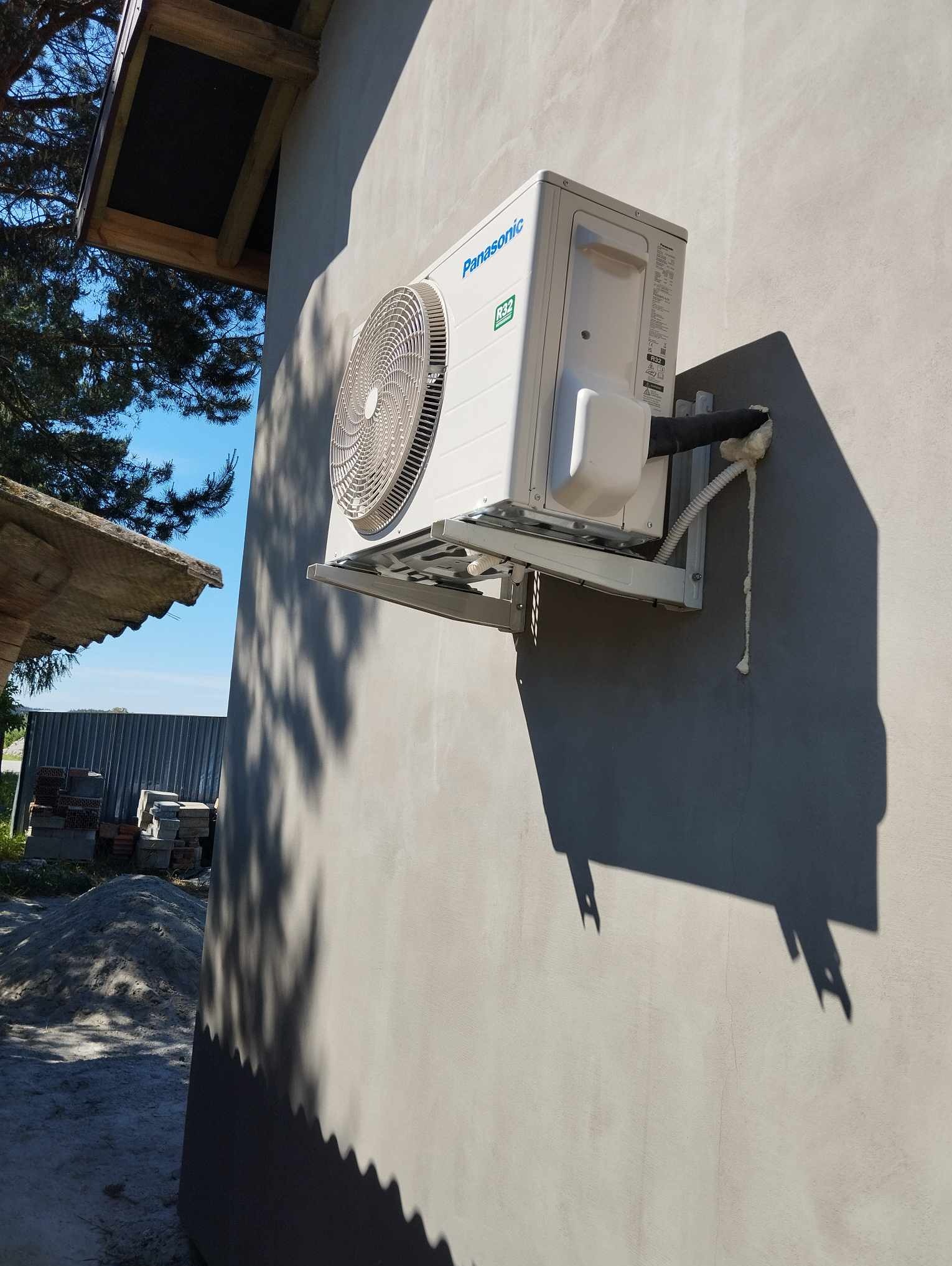 Zewnętrzna jednostka klimatyzacji Panasonic zamontowana na ścianie budynku, widoczne przewody zabezpieczone pianką montażową, w tle sterta cegieł i zarys dachu.