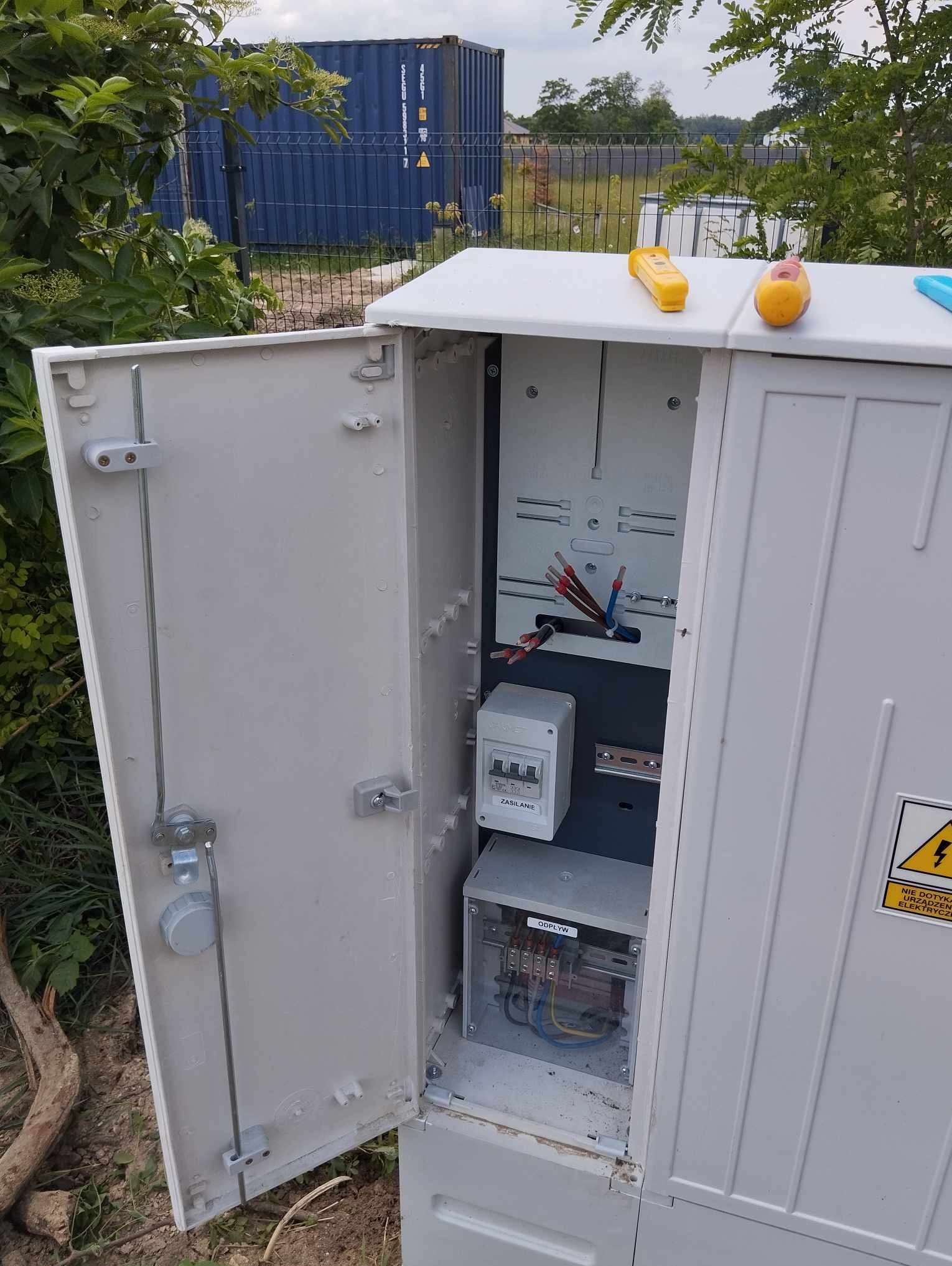 Otwarta skrzynka elektryczna z widocznymi przewodami, bezpiecznikami i oznaczeniami 'ZASILANIE' i 'ODPŁYW', umieszczona na zewnątrz, na tle zieleni i kontenera.