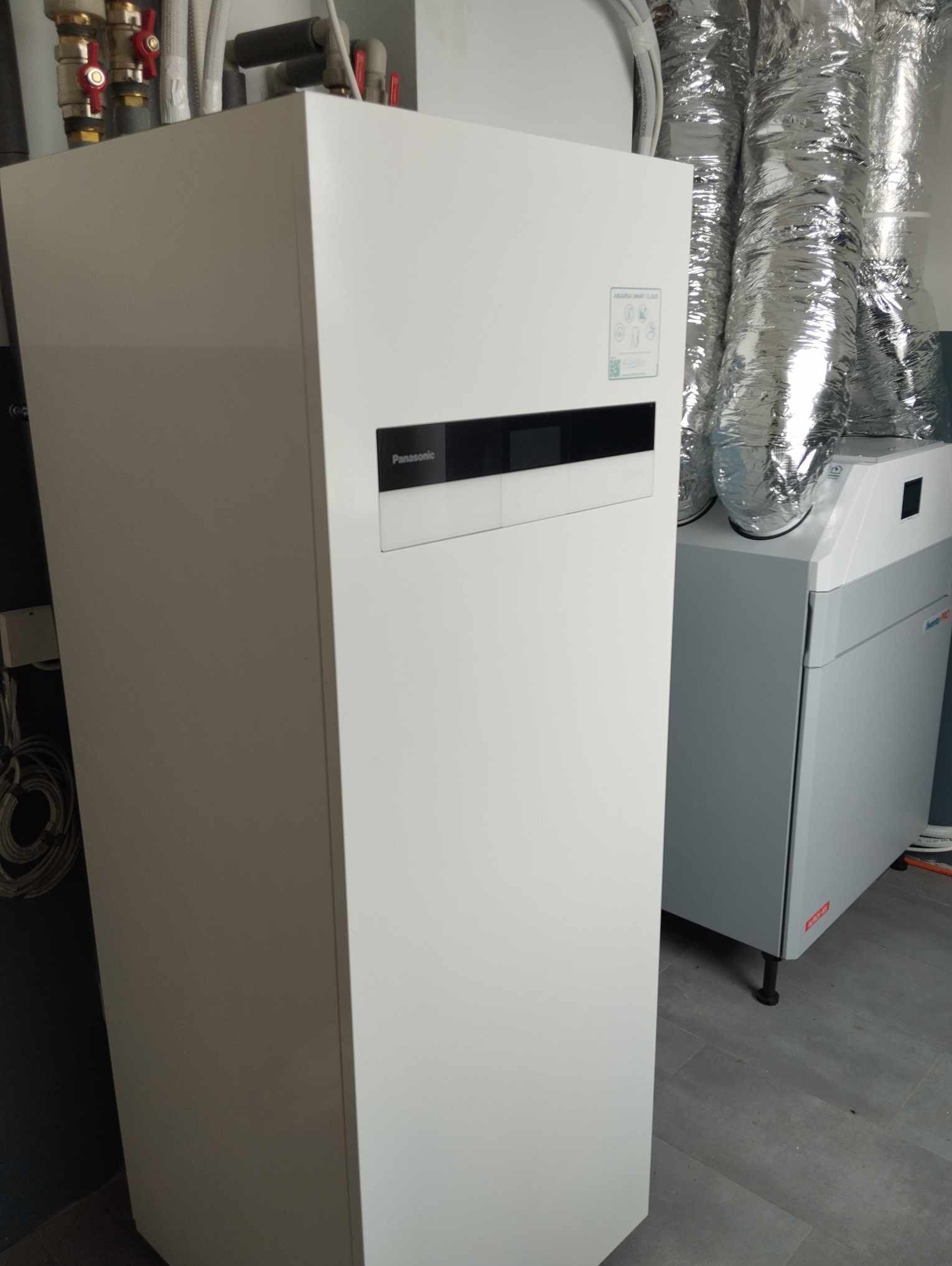 Biała pompa ciepła Panasonic Aquarea Smart Cloud z widocznymi rurami instalacyjnymi i jednostką wentylacyjną z metalowymi przewodami wentylacyjnymi w tle.