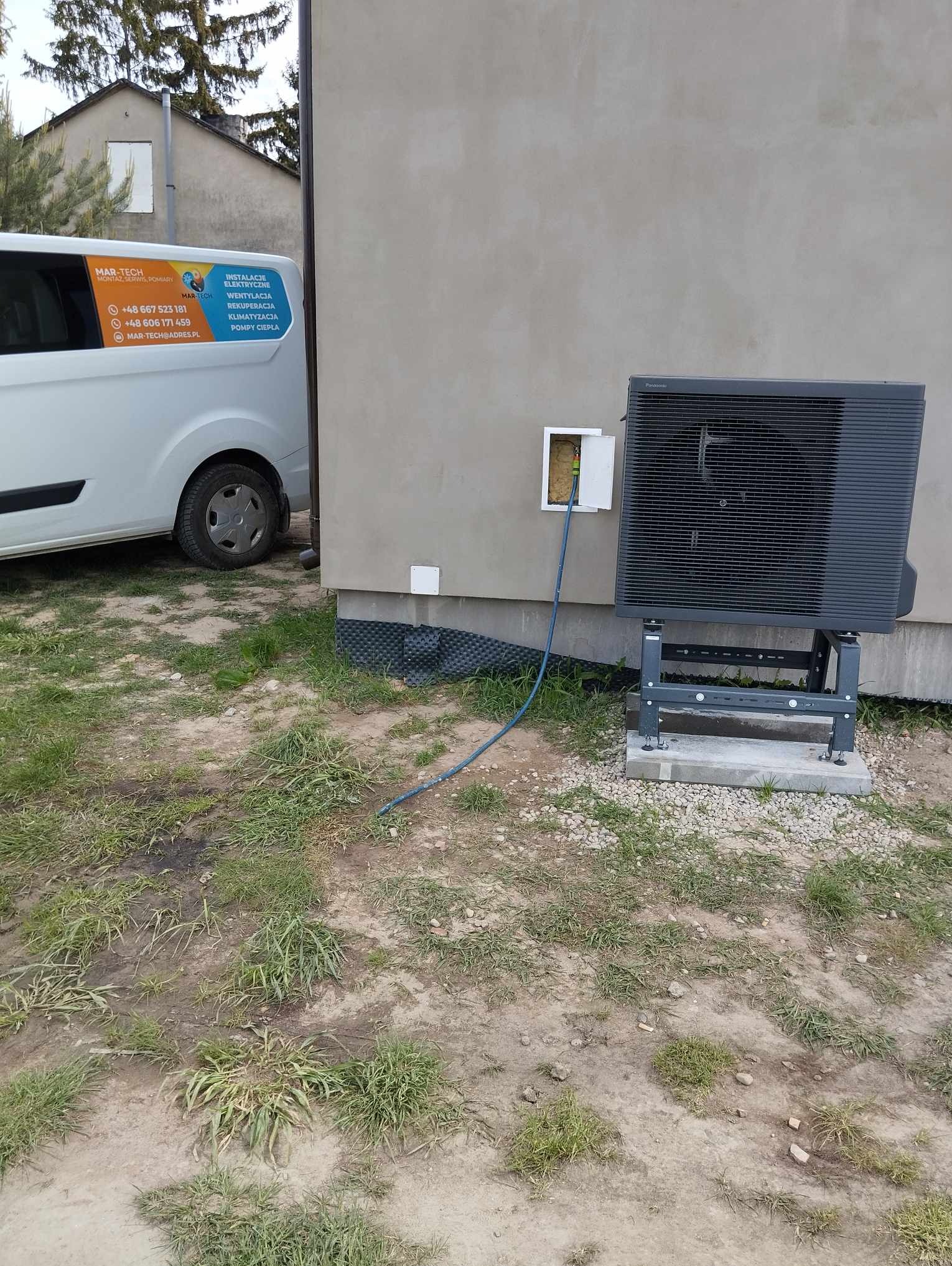 Czarna pompa ciepła zamontowana na szarym fundamencie obok ściany budynku, z widocznym kablem wchodzącym do skrzynki elektrycznej, na pierwszym planie trawa i częściowo widoczny samochód firmy...