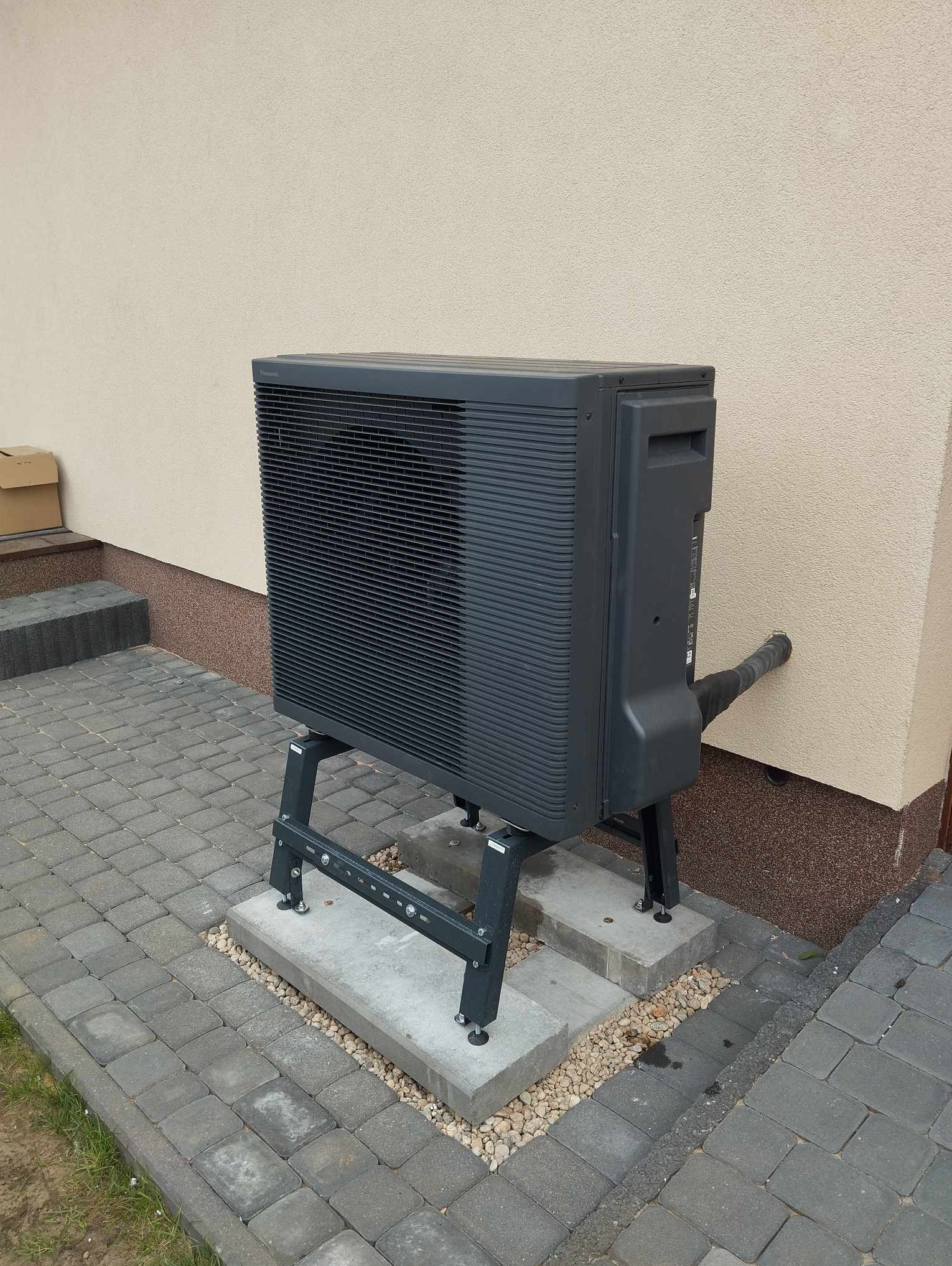 Szara zewnętrzna jednostka pompy ciepła zamontowana na metalowym stelażu, z rurą przechodzącą przez ścianę budynku, na tle kostki brukowej i elewacji.