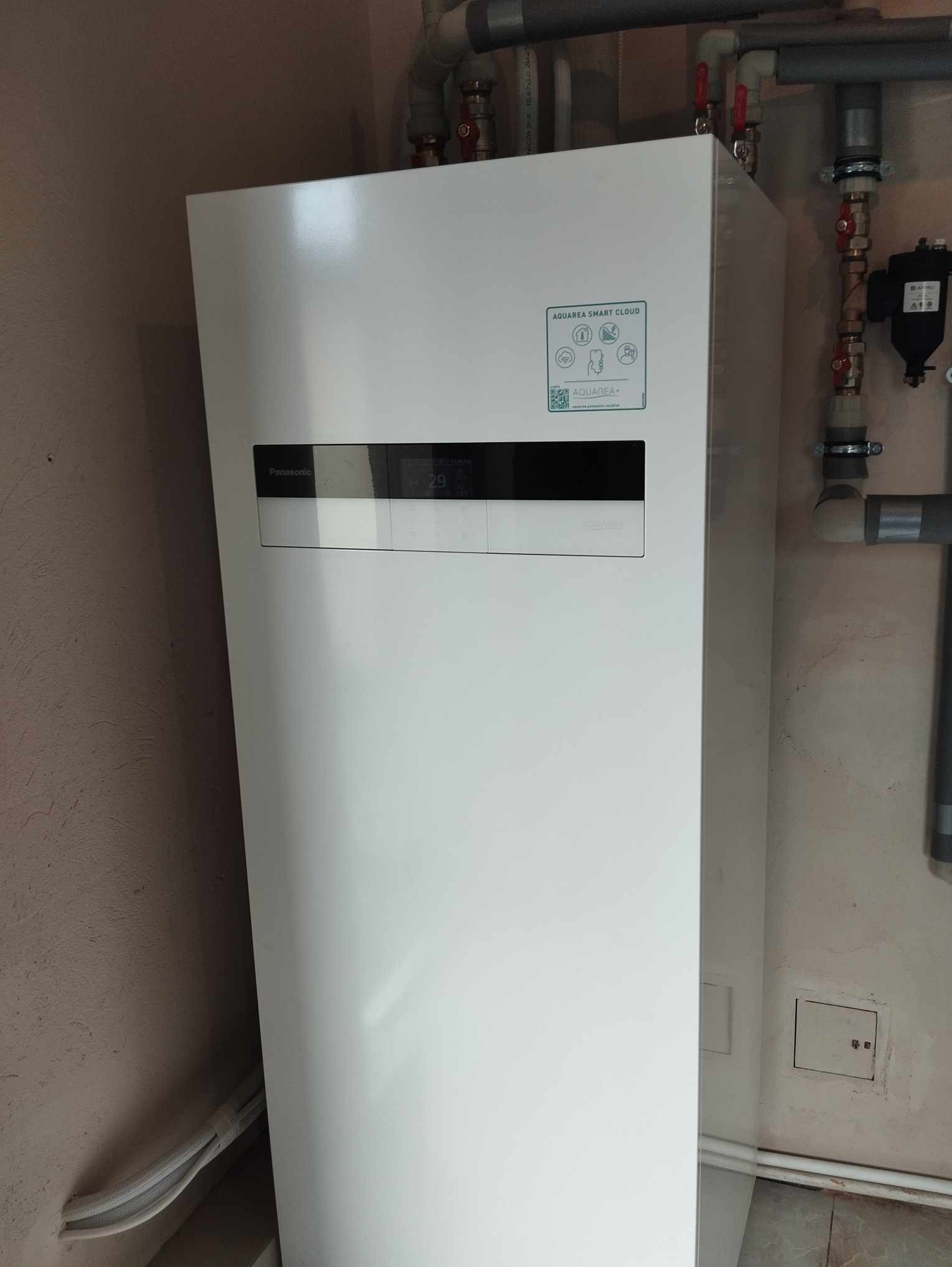 Biała pompa ciepła Panasonic Aquarea Smart Cloud zainstalowana w pomieszczeniu z widocznymi rurami i zaworami.