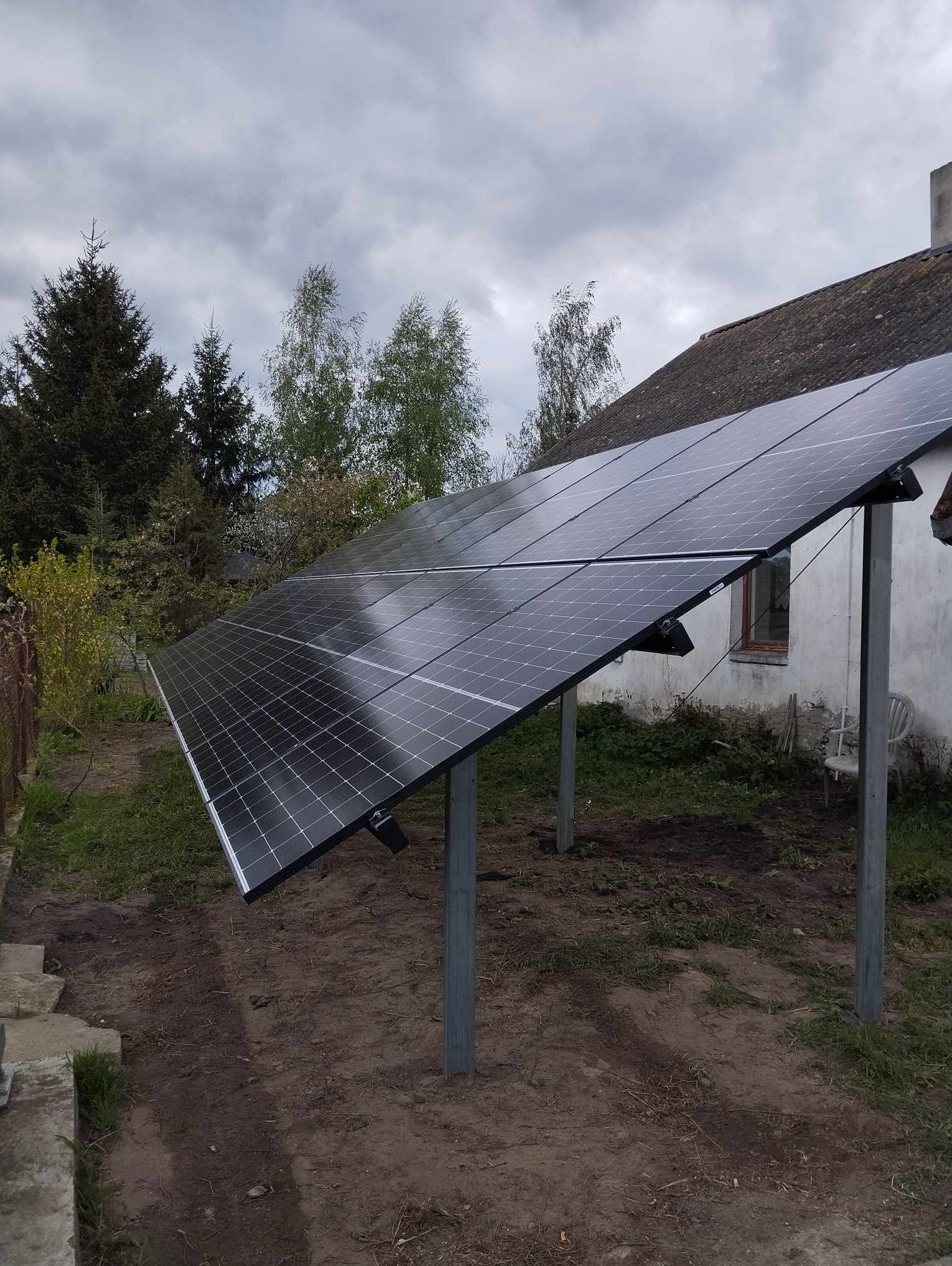 Zestaw paneli słonecznych zamontowany na metalowych słupach obok starego, białego domu z szarym dachem w pochmurny dzień.