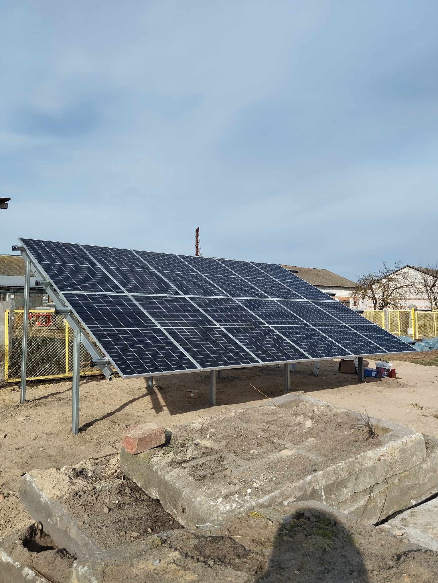 Panel fotowoltaiczny na metalowej konstrukcji, widok z dołu, w tle zabudowania gospodarcze i błękitne niebo z chmurami, na pierwszym planie fragmenty betonowych płyt.