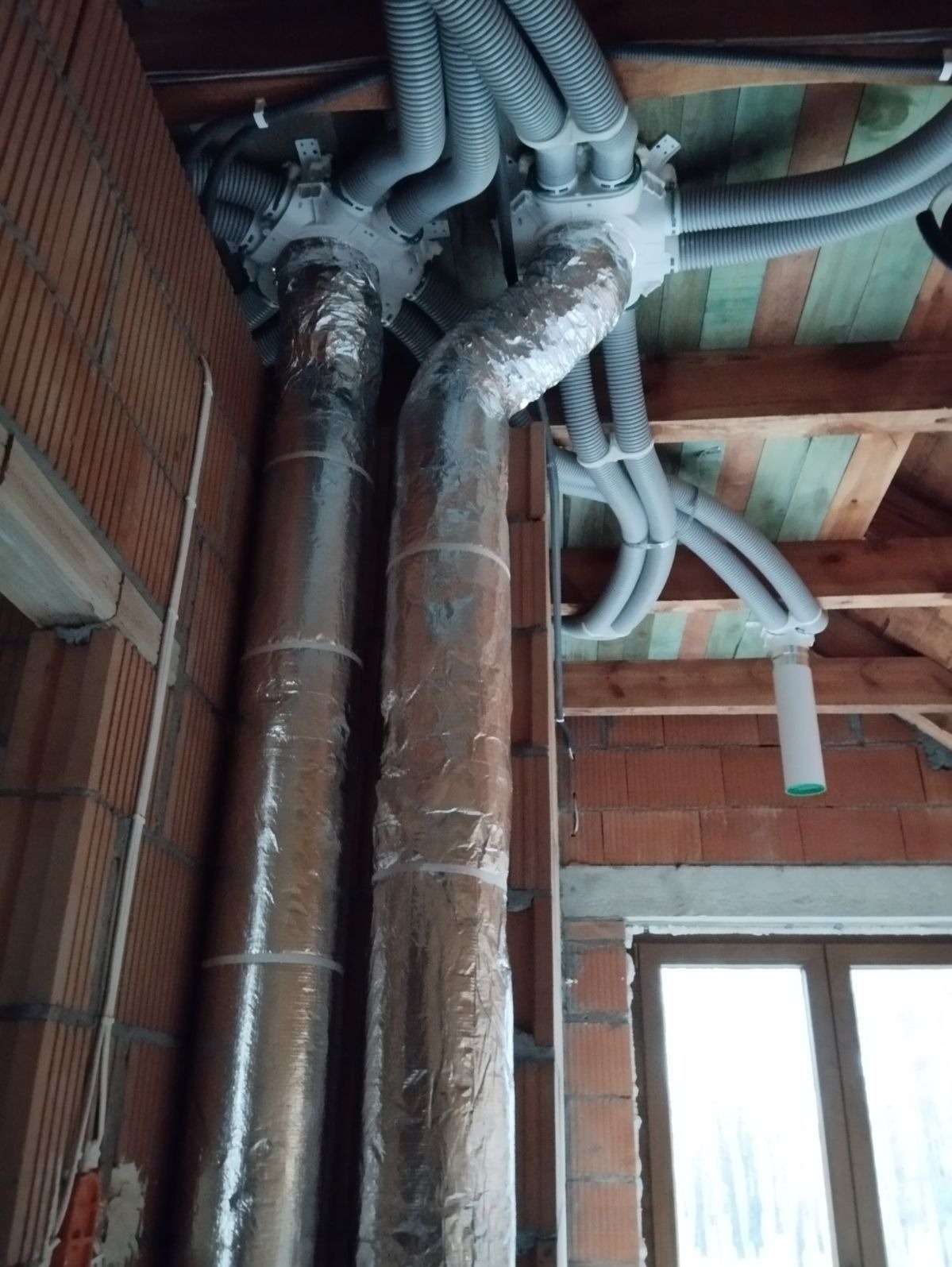Dwie pionowe rury owinięte folią aluminiową, połączone z szarymi elastycznymi przewodami wentylacyjnymi i białymi rozdzielaczami w surowym wnętrzu poddasza z ceglanymi ścianami i drewnianą...