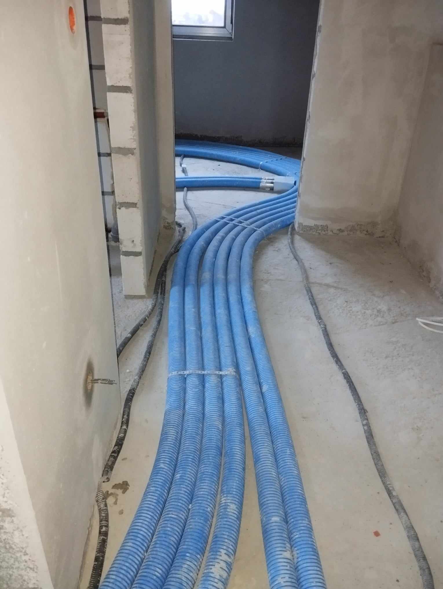 Niebieskie rury rekuperacji ułożone w rzędach na surowym betonowym podłożu w trakcie montażu w nowym budynku, widoczne przewody elektryczne w peszlach.