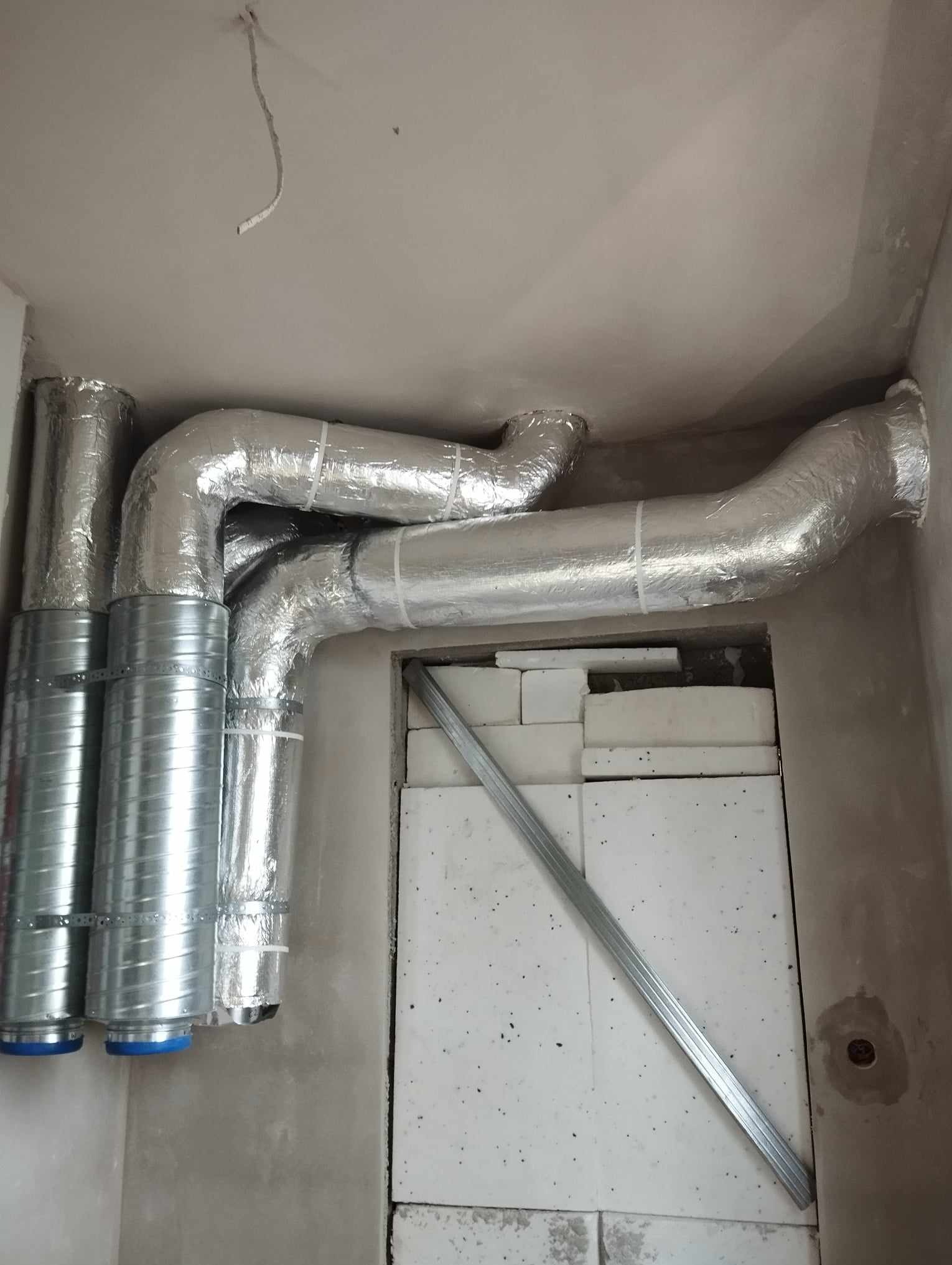 System rur wentylacyjnych, częściowo owiniętych folią aluminiową, zamontowany pod sufitem w surowym wnętrzu. Widoczne elementy mocujące i białe płyty izolacyjne w otworze ściany.