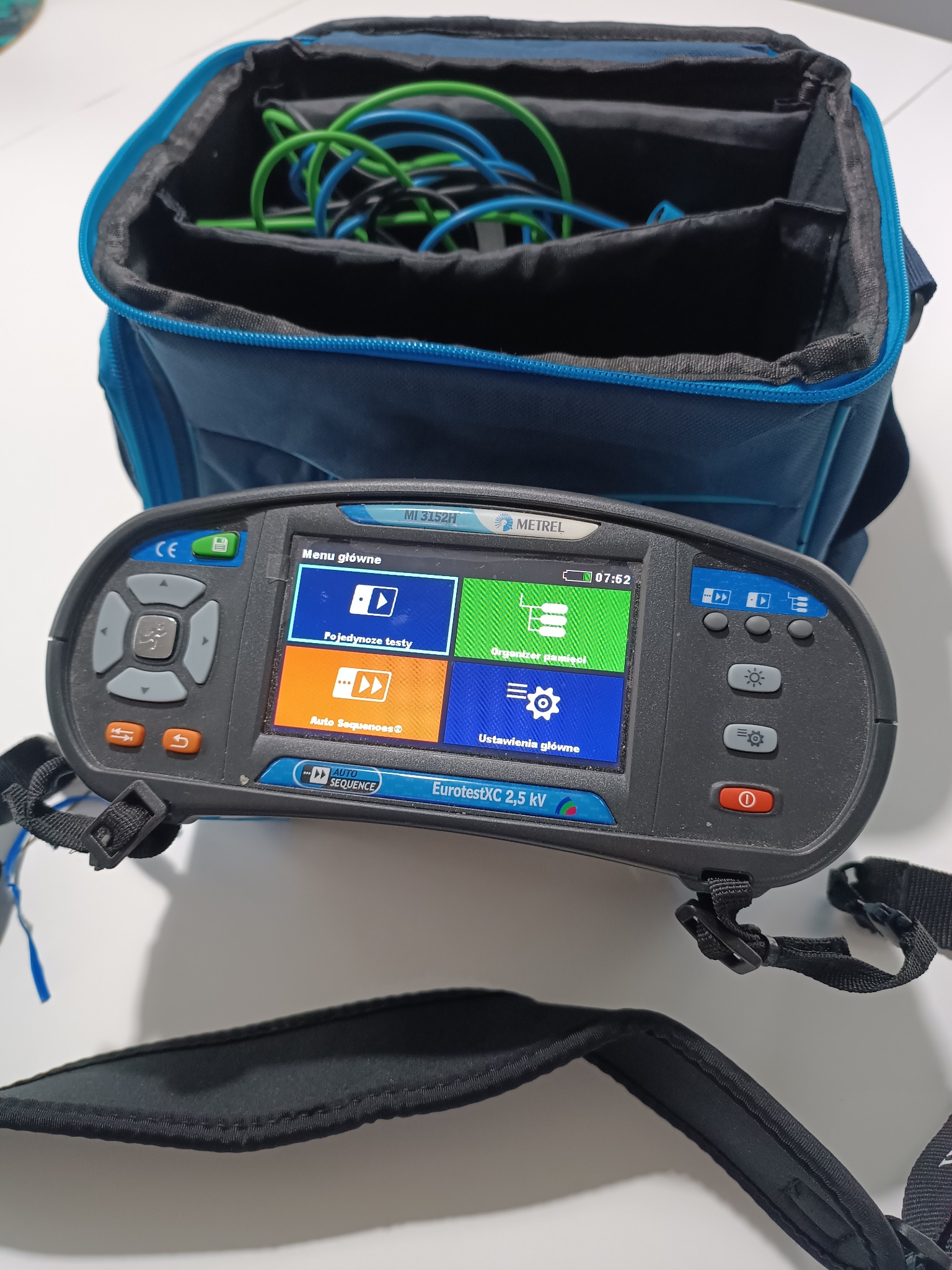 Miernik parametrów instalacji elektrycznej EurotestXC 2,5 kV firmy Metrel leżący na torbie z przewodami pomiarowymi w kolorach zielonym i niebieskim.