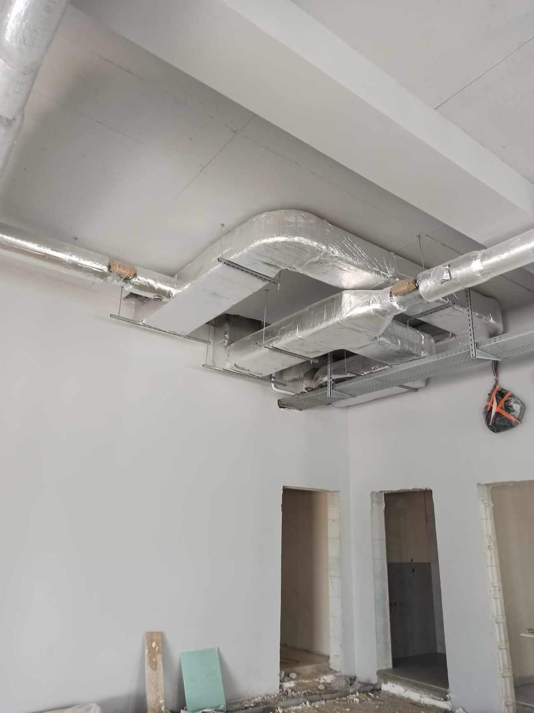 Wnętrze w trakcie wykańczania z widocznymi kanałami wentylacyjnymi pod sufitem, zabezpieczonymi folią, oraz przygotowanymi otworami drzwiowymi.