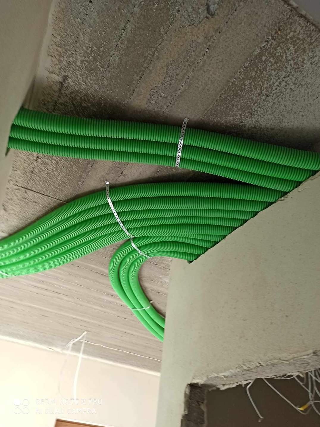 Zielone rury karbowane przymocowane metalowymi obejmami do sufitu z surowego betonu, widok instalacji w trakcie montażu.