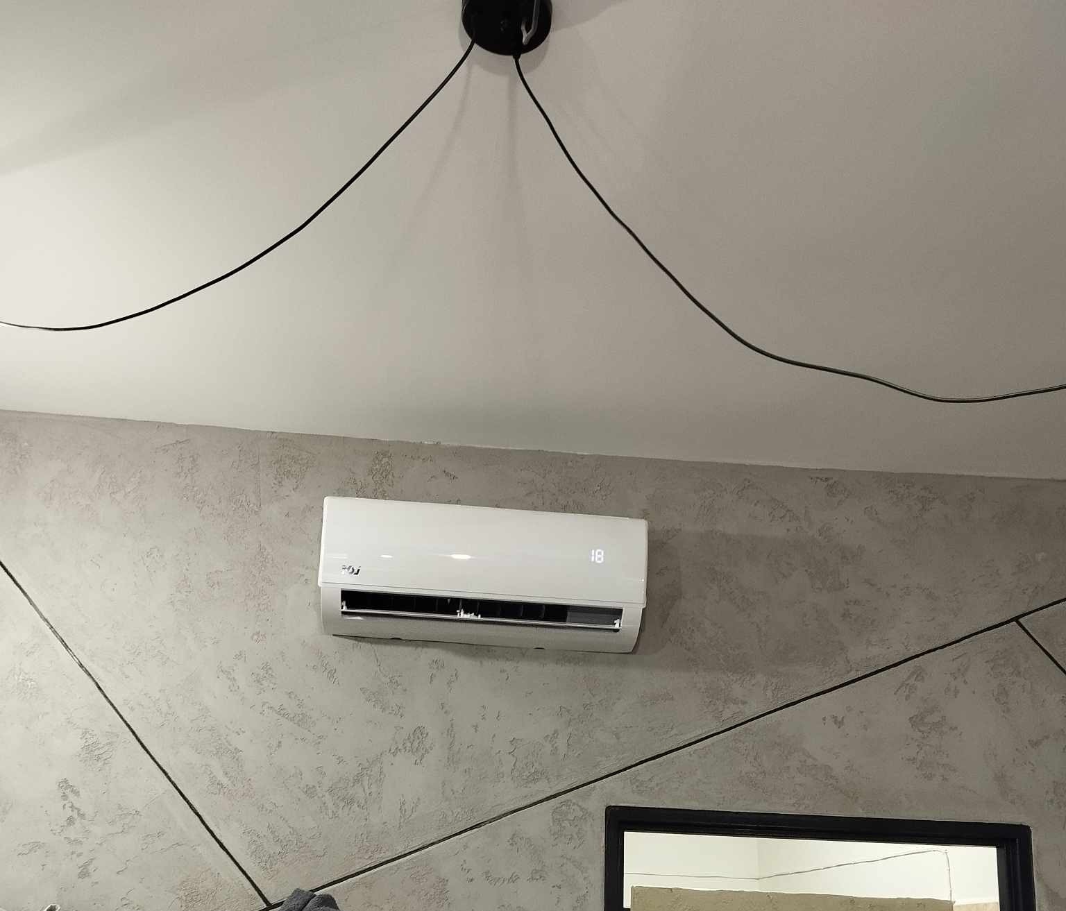 Biały klimatyzator ścienny z wyświetlaczem temperatury 18 stopni, zamontowany na szarej ścianie z dekoracyjnym wzorem geometrycznym, widoczne czarne kable od oświetlenia sufitowego.