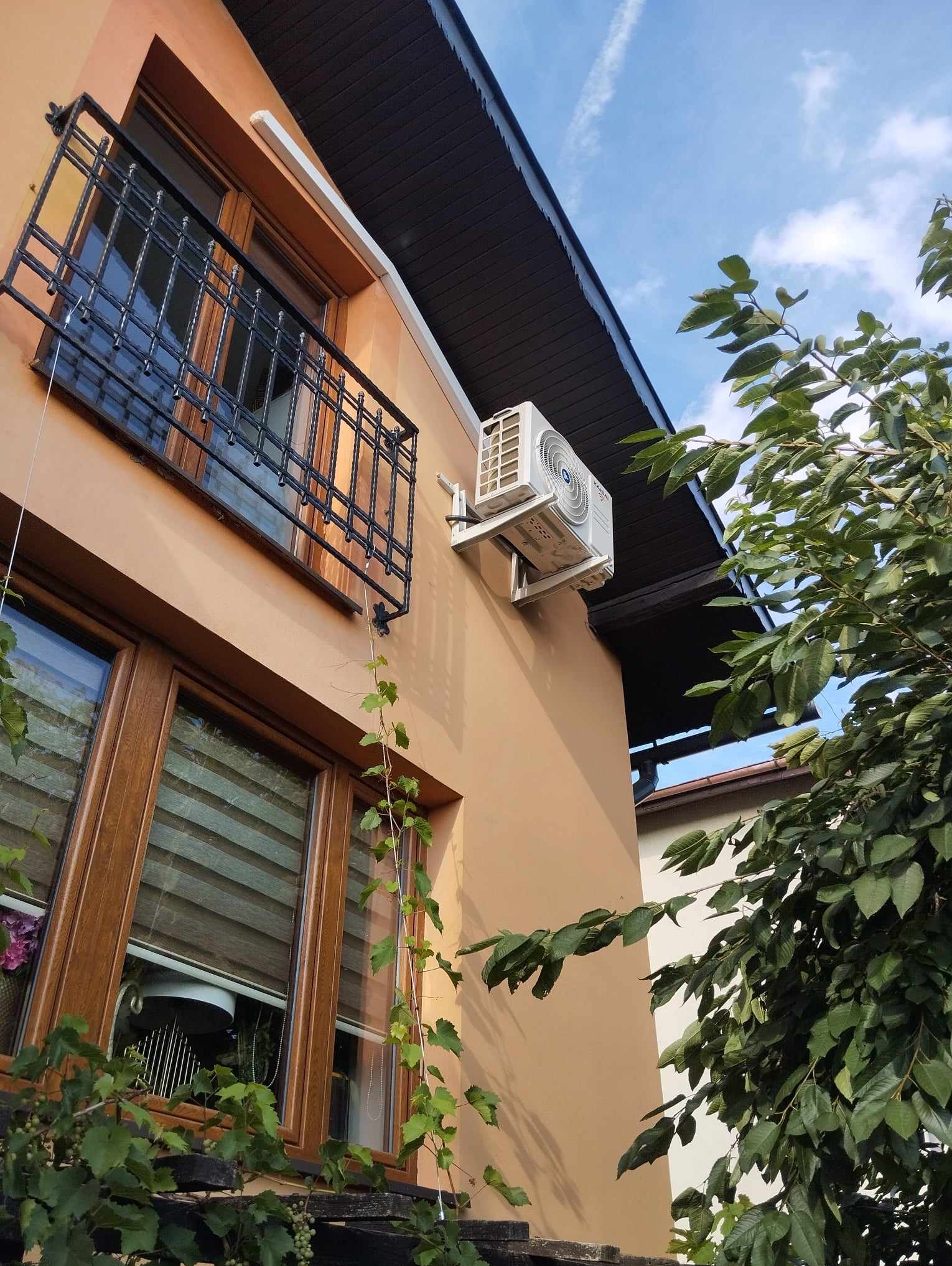 Biała jednostka zewnętrzna klimatyzatora zamontowana na elewacji budynku z oknami, widok z dołu, częściowo zasłonięta przez gałęzie drzewa.