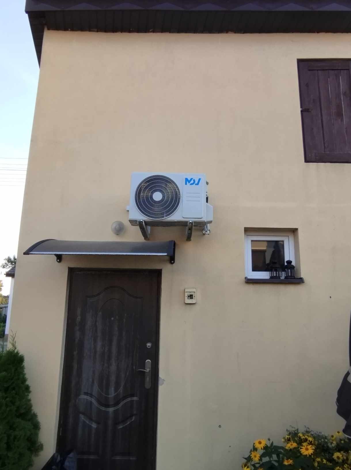 Zewnętrzna jednostka klimatyzacji zamontowana na elewacji domu, widoczna nad wejściem z zadaszeniem i małym oknem z ozdobami.