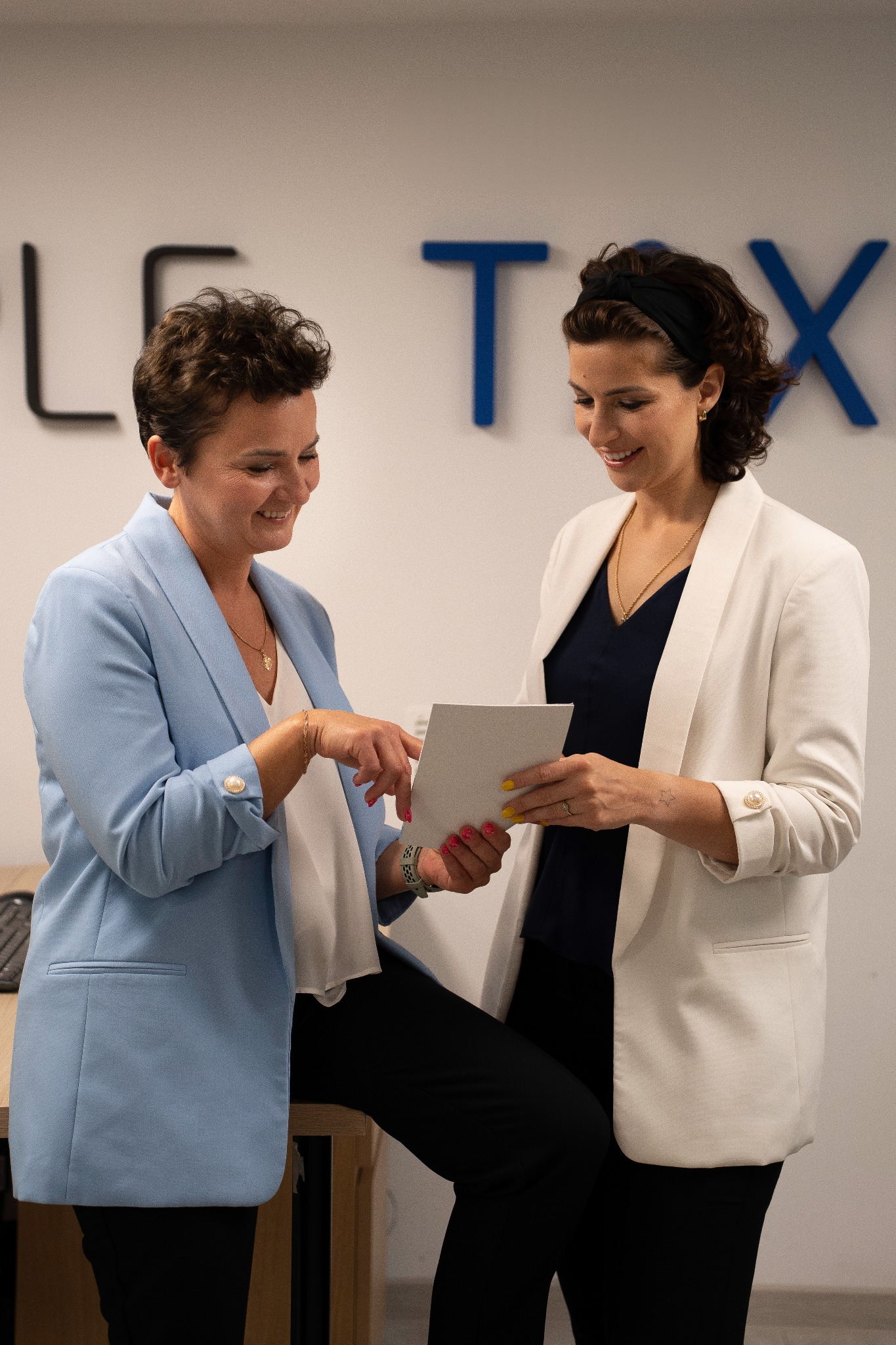 Dwie uśmiechnięte kobiety w biurowych strojach analizują dokument, na ścianie widoczny napis z niebieskimi literami.