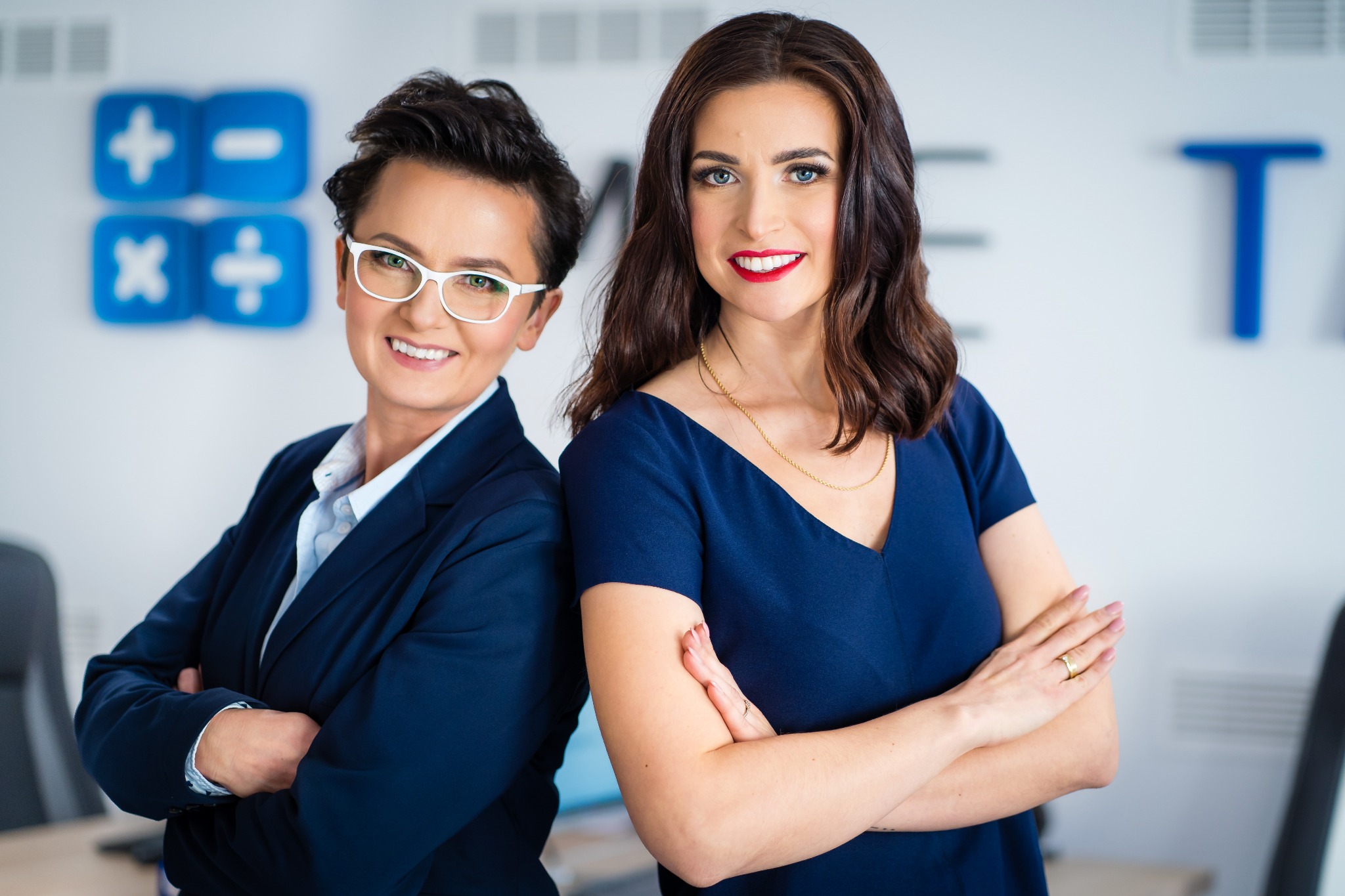 Dwie uśmiechnięte kobiety w biurowym otoczeniu, stojące plecami do siebie, jedna w okularach i granatowym garniturze, druga w granatowej sukience, w tle symbole matematyczne i litery.