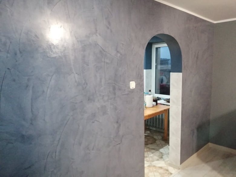 Szara ściana z efektem dekoracyjnym, widoczny łuk wejściowy do kuchni z oknem. Wykończenie wnętrza w odcieniach szarości, minimalistyczny styl.