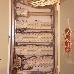 Jakub Zblewski - Rozdzielnica elektryczna w trakcie montażu, widoczne liczne przewody i zabezpieczenia, puszki instalacyjne na ścianie gipsowej, etap prac instalacyjnych.