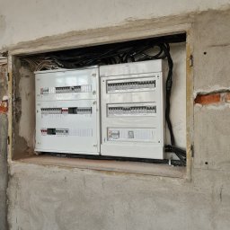 Jakub Zblewski - Dwie białe skrzynki rozdzielcze z bezpiecznikami, zamontowane w surowej wnęce w ścianie z widocznymi przewodami elektrycznymi.