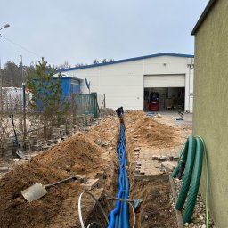 Jakub Zblewski - Wykop z ułożonymi niebieskimi rurami karbowanymi, widoczny pracownik w oddali, hałda piasku, kostka brukowa, budynek magazynowy w tle, pochmurny dzień.