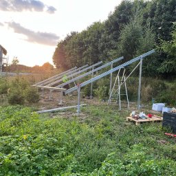 Jakub Zblewski - Montaż konstrukcji wsporczej pod panele słoneczne na działce z widokiem na dom w stanie surowym, narzędzia na palecie i drabiny w tle.