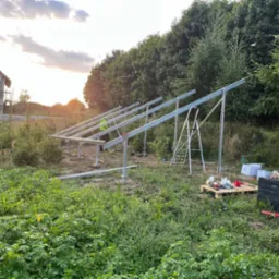 Montaż konstrukcji wsporczej pod panele słoneczne na działce z widokiem na dom w stanie surowym, narzędzia na palecie i drabiny w tle.