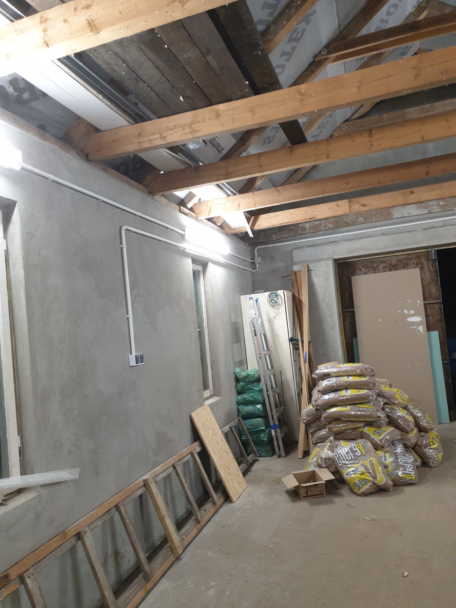 Wnętrze remontowanego pomieszczenia z surowymi ścianami, widoczną instalacją elektryczną w białych rurkach, drewnianą konstrukcją dachu i stertą worków z pelletem w rogu.