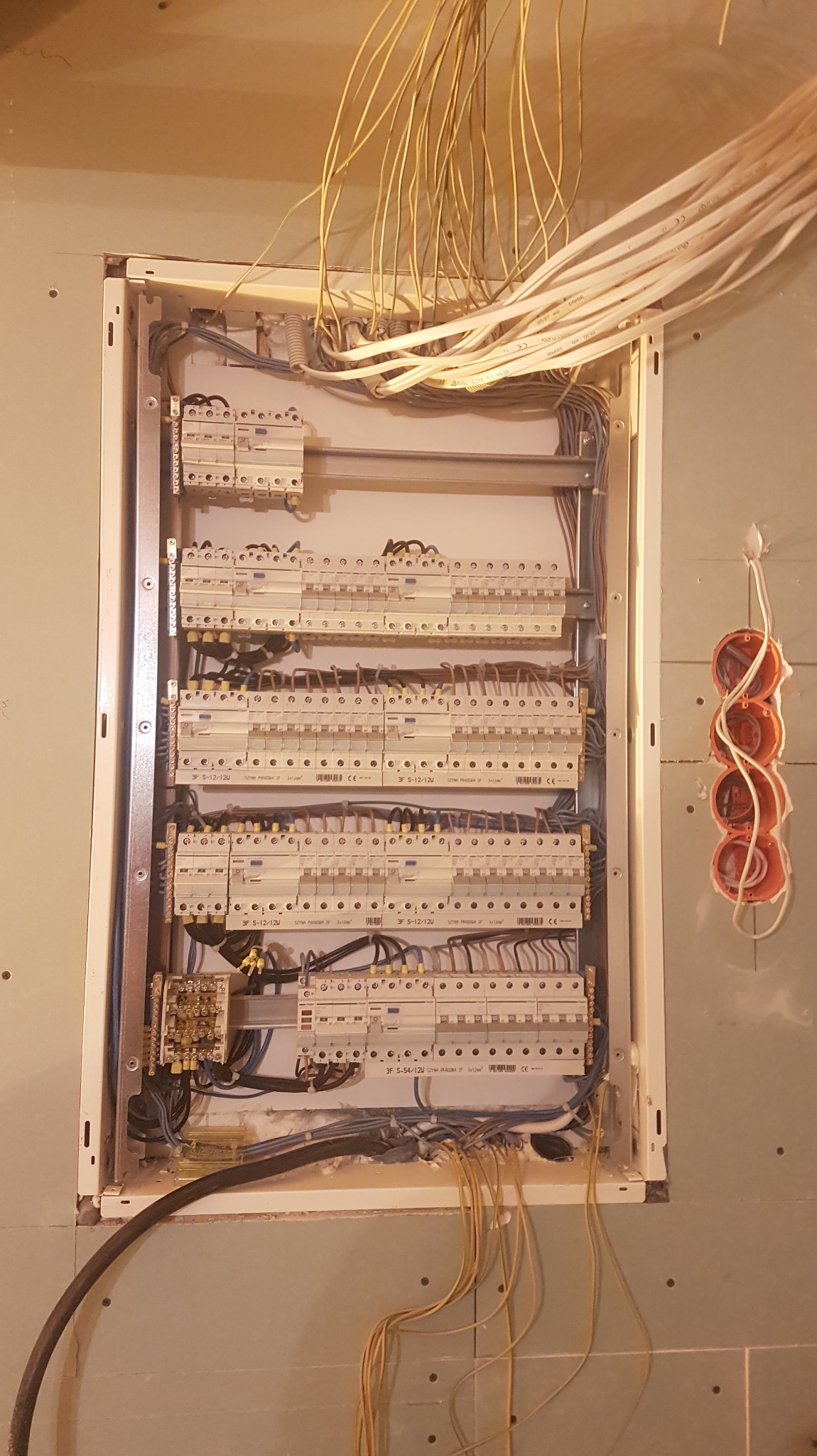 Rozdzielnica elektryczna w trakcie montażu, widoczne liczne przewody i zabezpieczenia, puszki instalacyjne na ścianie gipsowej, etap prac instalacyjnych.