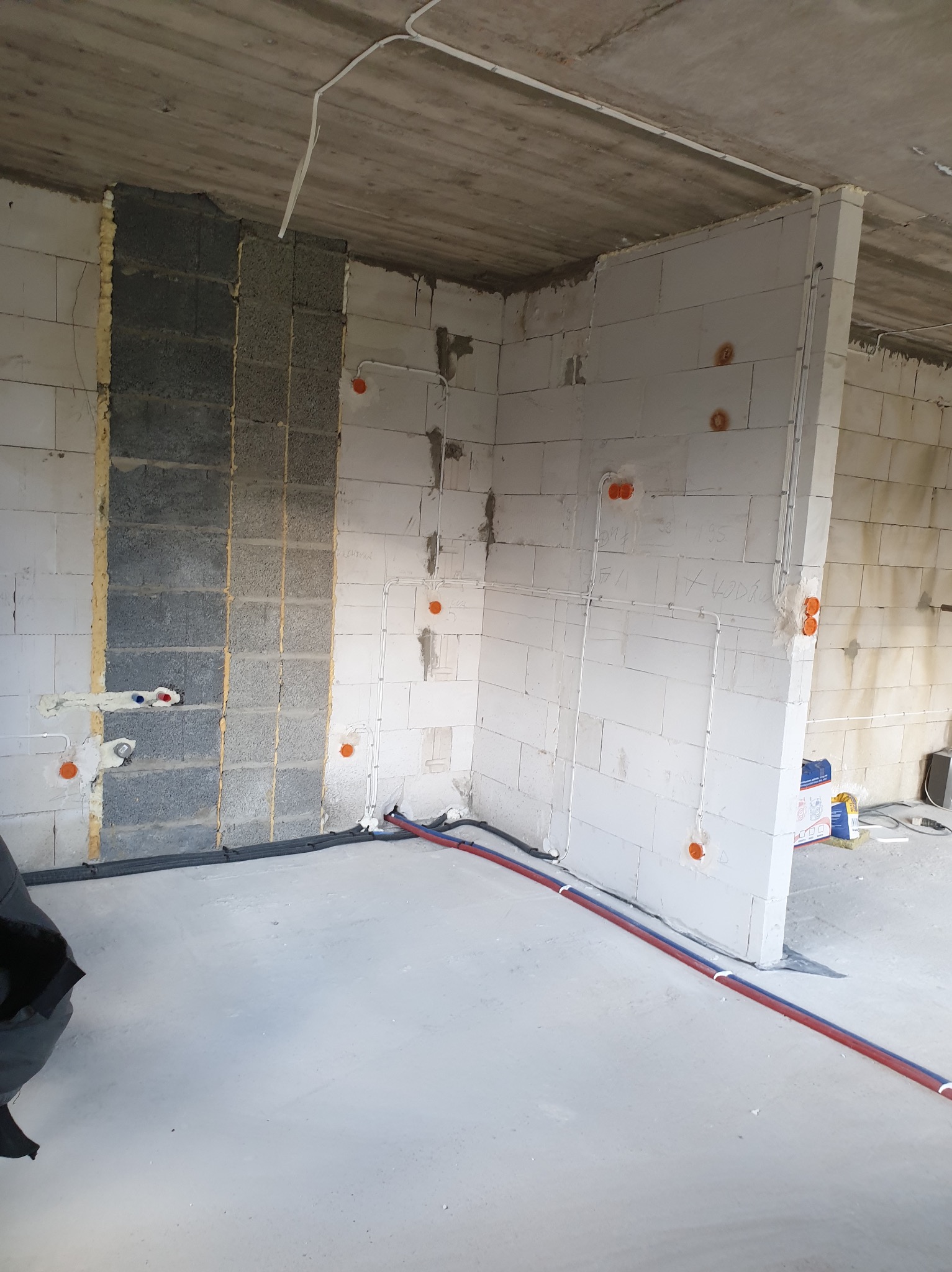 Surowy stan wnętrza budynku w trakcie prac instalacyjnych: widoczne przewody elektryczne w peszlach na ścianach z betonu komórkowego, rury instalacji wodnej w posadzce, puszki elektryczne. Fragment...