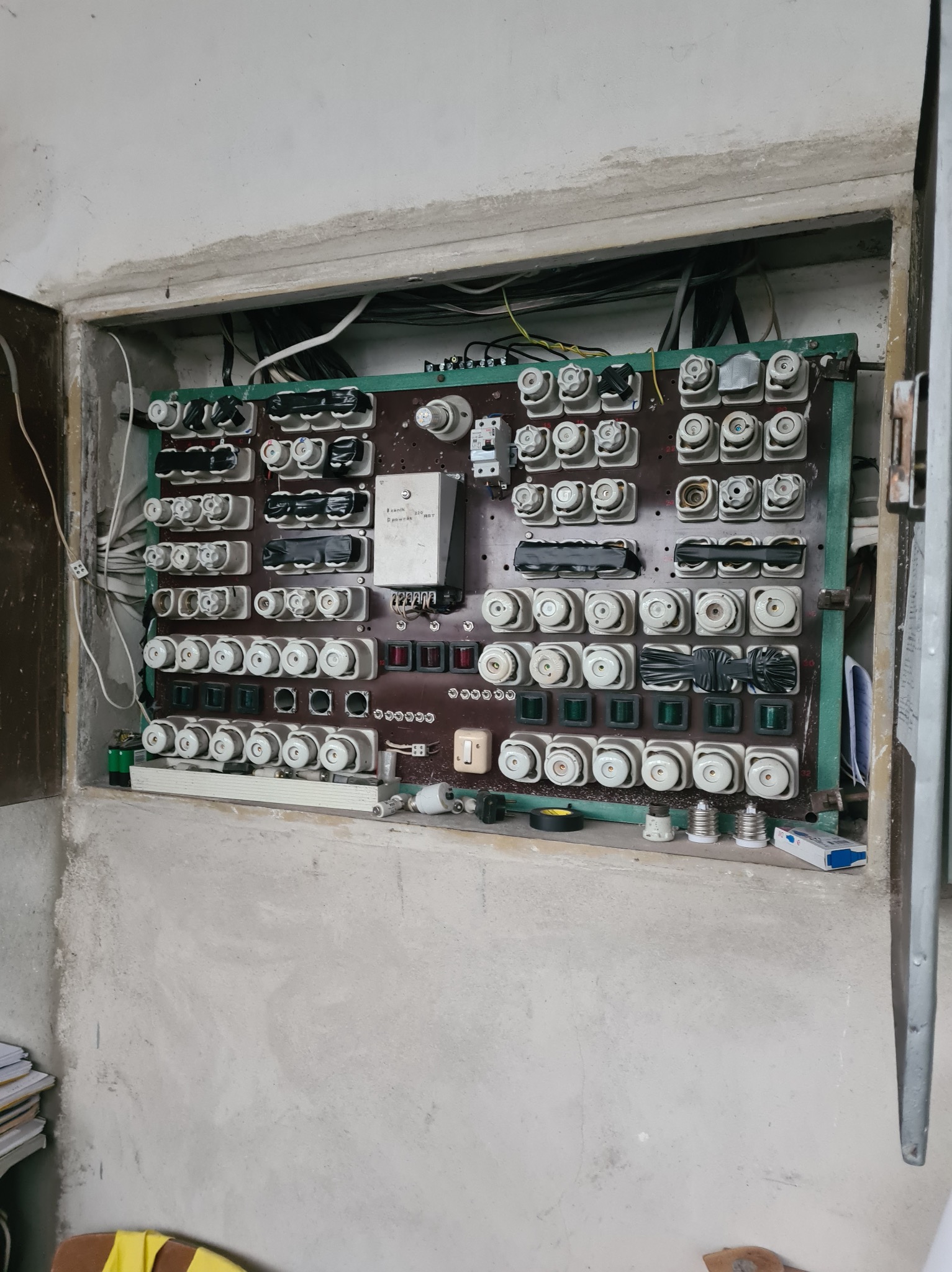 Stara, brązowa tablica rozdzielcza z bezpiecznikami topikowymi, umieszczona w szarej, betonowej wnęce. Widoczne kable, przewody i elementy instalacji elektrycznej. Niektóre bezpieczniki zaklejone...