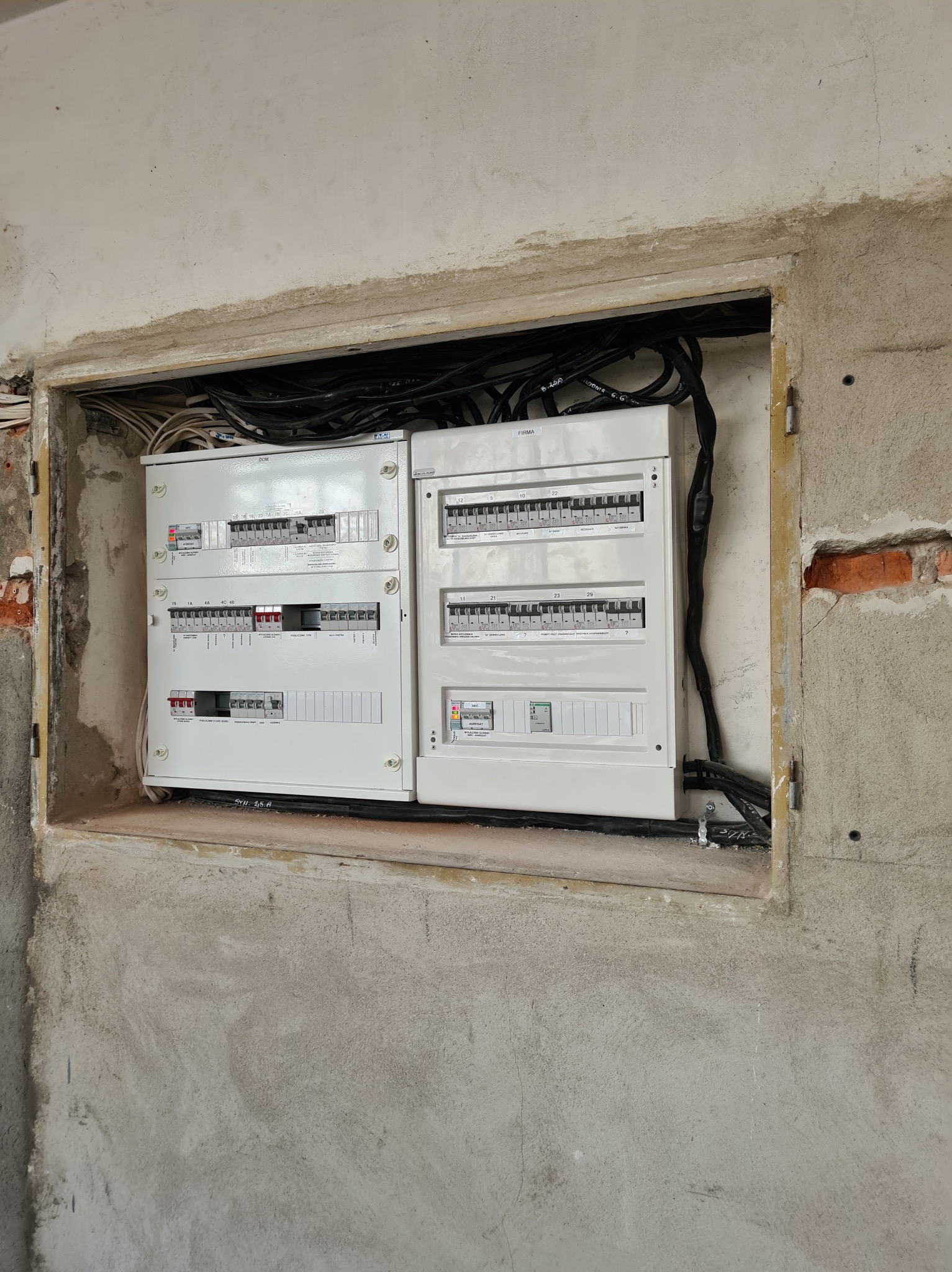 Dwie białe skrzynki rozdzielcze z bezpiecznikami, zamontowane w surowej wnęce w ścianie z widocznymi przewodami elektrycznymi.
