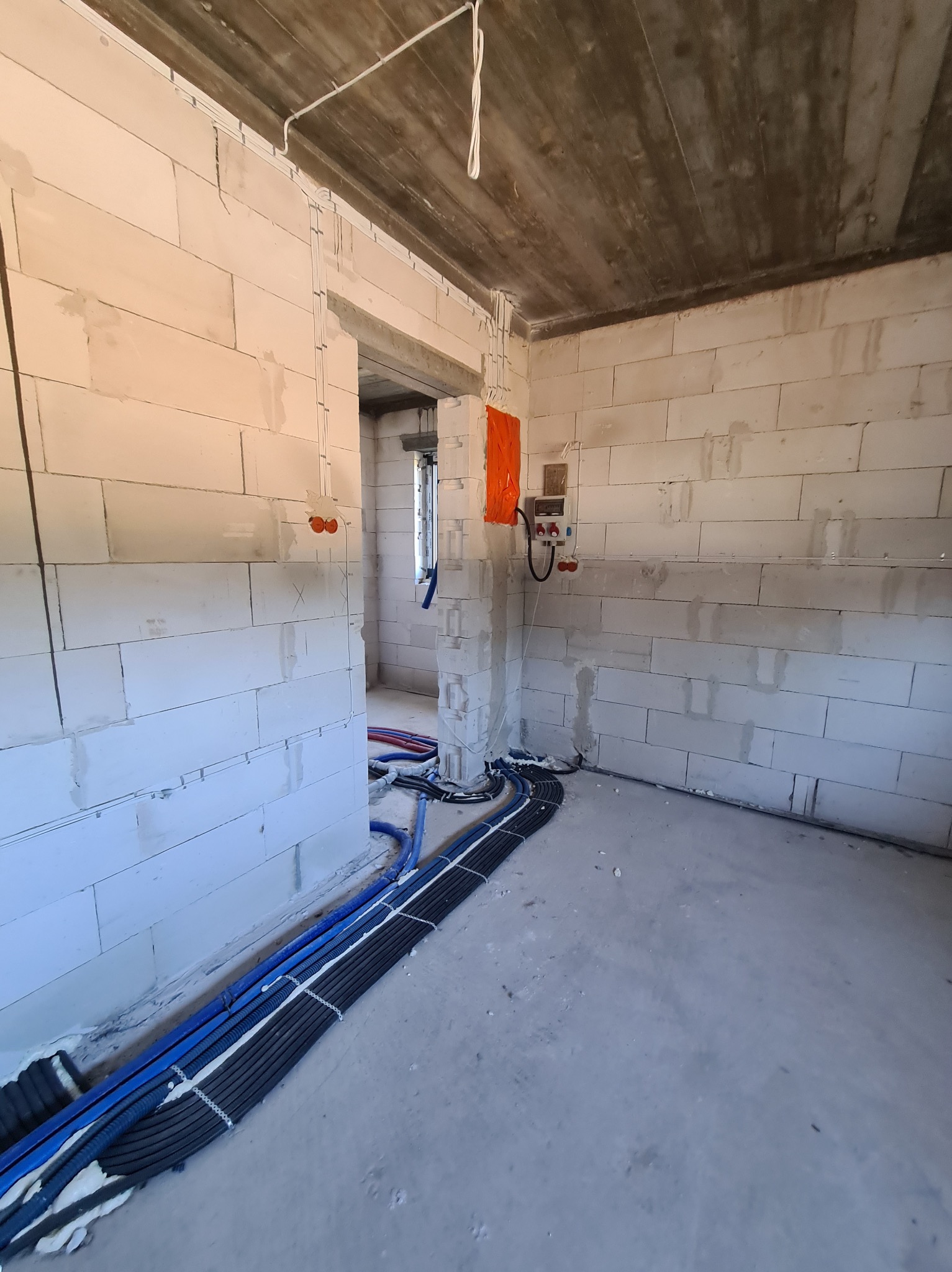 Wnętrze surowego budynku z widocznymi przewodami elektrycznymi i rurami instalacyjnymi ułożonymi wzdłuż ściany, skrzynka elektryczna z zabezpieczeniem pomarańczową folią, ściany z betonu...