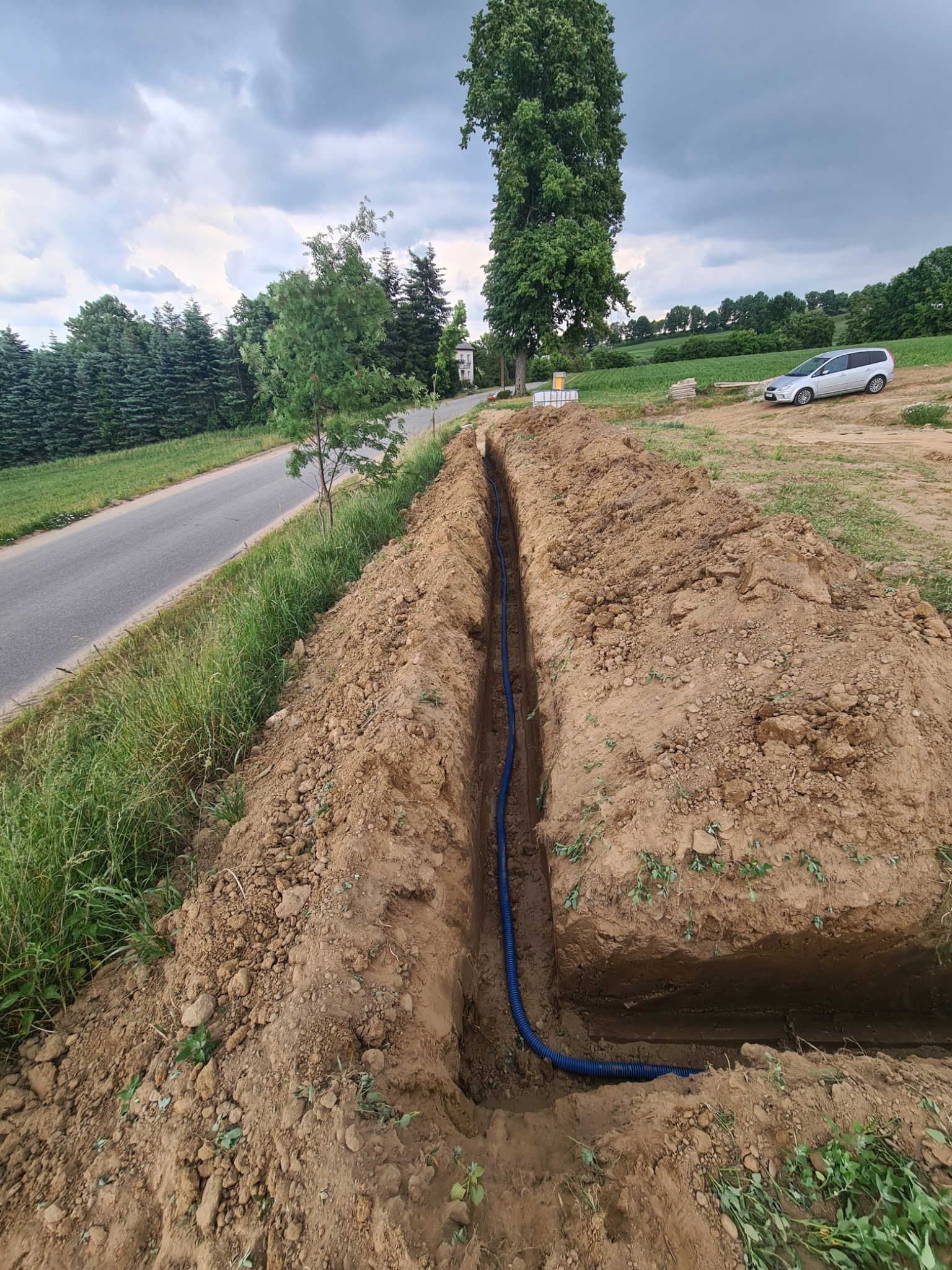 Wąski wykop w ziemi z ułożoną niebieską rurą karbowaną, prawdopodobnie pod kabel elektryczny, widok na drogę asfaltową, drzewa i pole w tle.